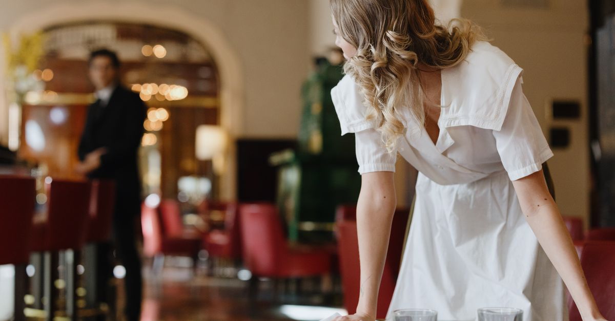 A restaurant waitress in white dress arranging tableware indoors, showcasing hospitality.