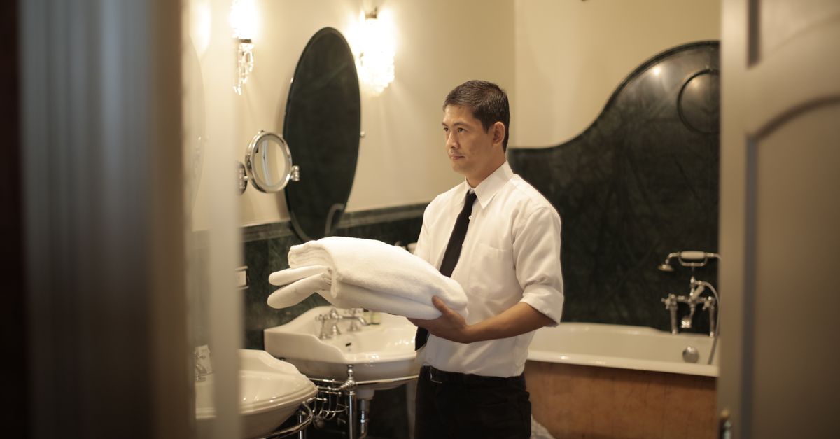 Side view of young male employee holding stack of white towels while standing in bathroom of luxury hotel