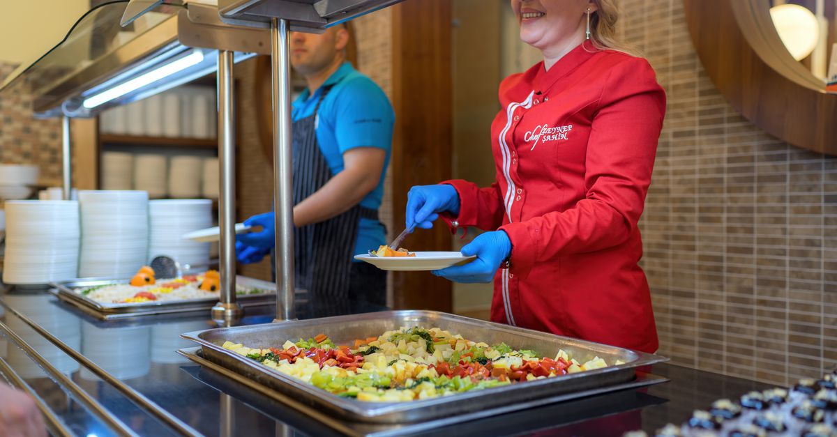 Chef in red uniform serving food at a buffet counter in Antalya restaurant, smiling and engaging with customers.