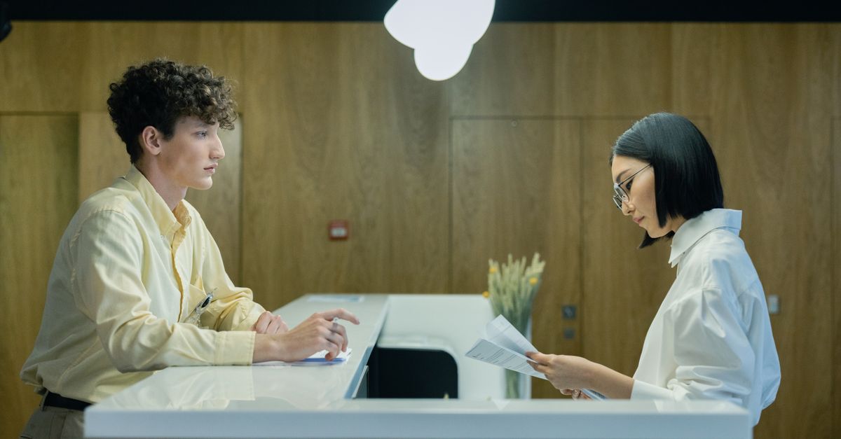 A young man and woman interacting at a modern office reception desk indoors.
