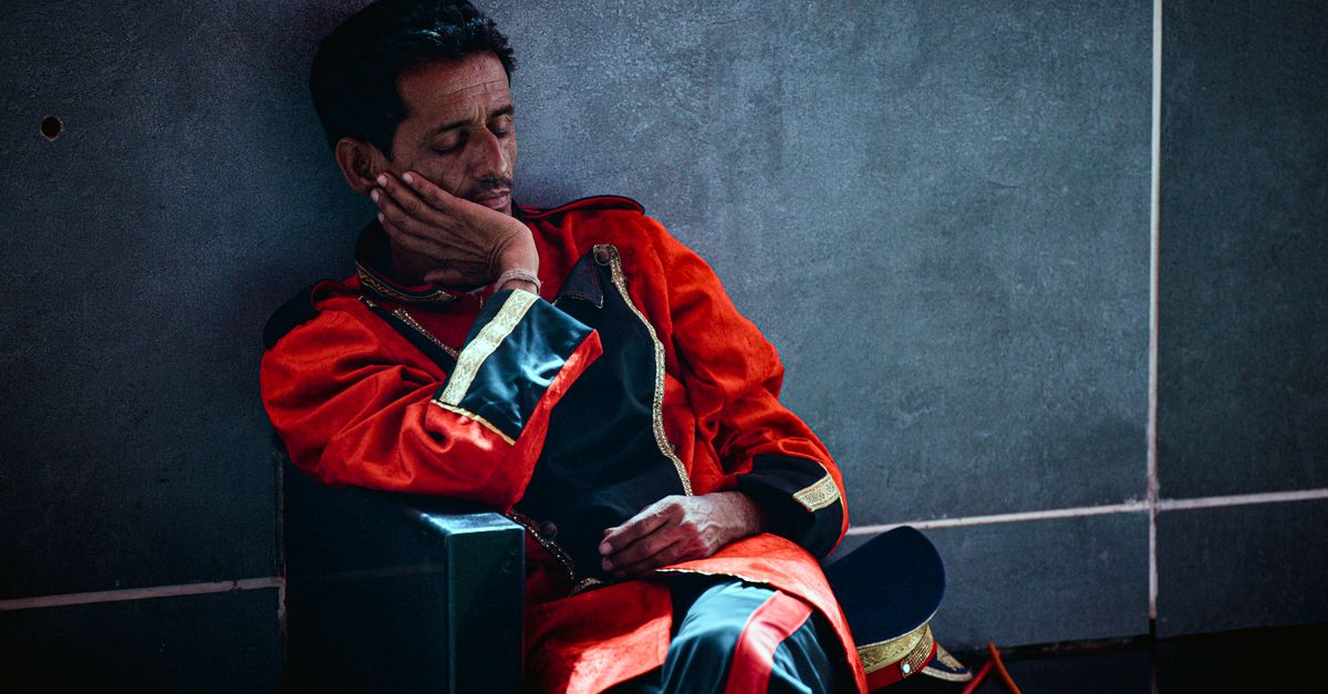 A hotel bellman in red uniform sleeps on a bench indoors, depicting exhaustion and rest.