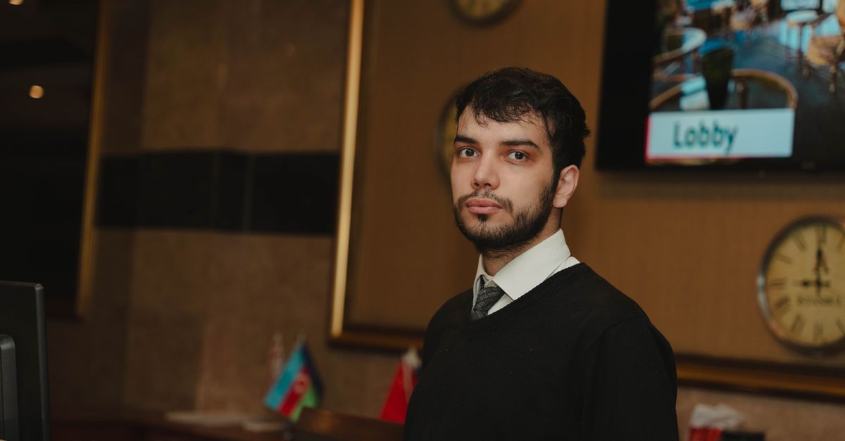 A hotel receptionist standing at the front desk in a hotel lobby, ready to assist guests.
