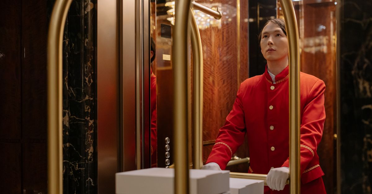 A hotel porter in a red uniform pushes a luggage cart in a luxurious elevator.
