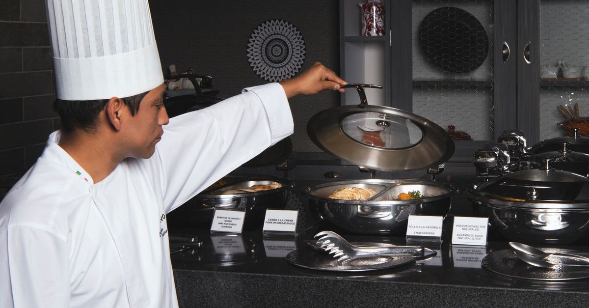 Chef in white uniform opening lid of buffet dish in modern kitchen setting.