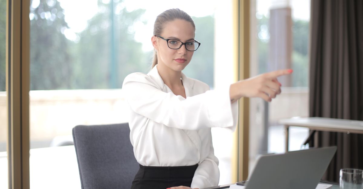 Concentrated businesswoman wearing formal clothes and eyeglasses pointing away while standing in office and working on project in laptop during work