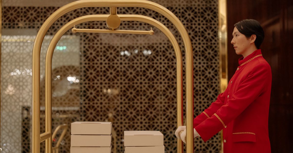 A hotel bellboy in uniform pushes a luggage cart with boxes indoors, demonstrating hospitality service.