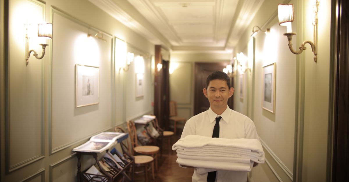 A young male hotel staff member carrying fresh towels in a beautifully lit hotel hallway.