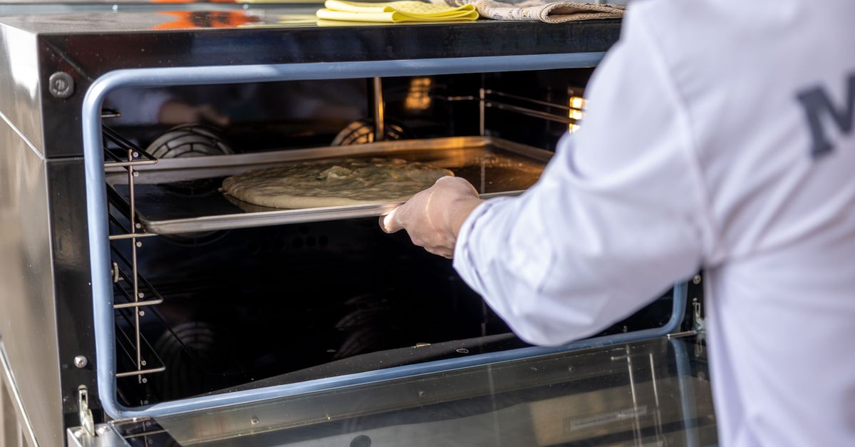 Chef places uncooked pizza into an industrial oven in a professional kitchen setting.